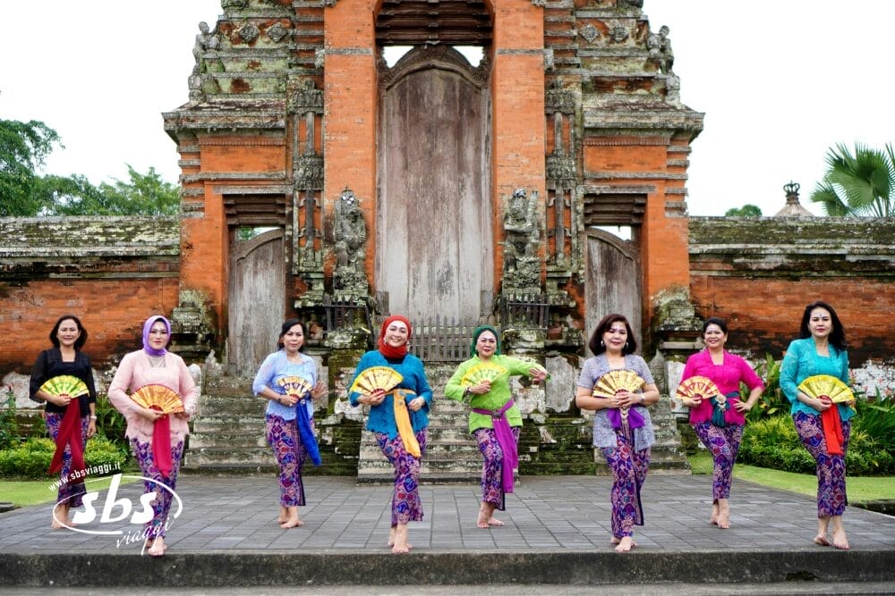 Otto donne in abiti tradizionali colorati tengono in mano dei ventagli e posano davanti al cancello di un tempio balinese ornato, con una vegetazione lussureggiante alle loro spalle. Il logo della SBS appare nell'angolo in basso a sinistra, evocando lo spirito vibrante di Bali e della vicina Gili Trawangan.
