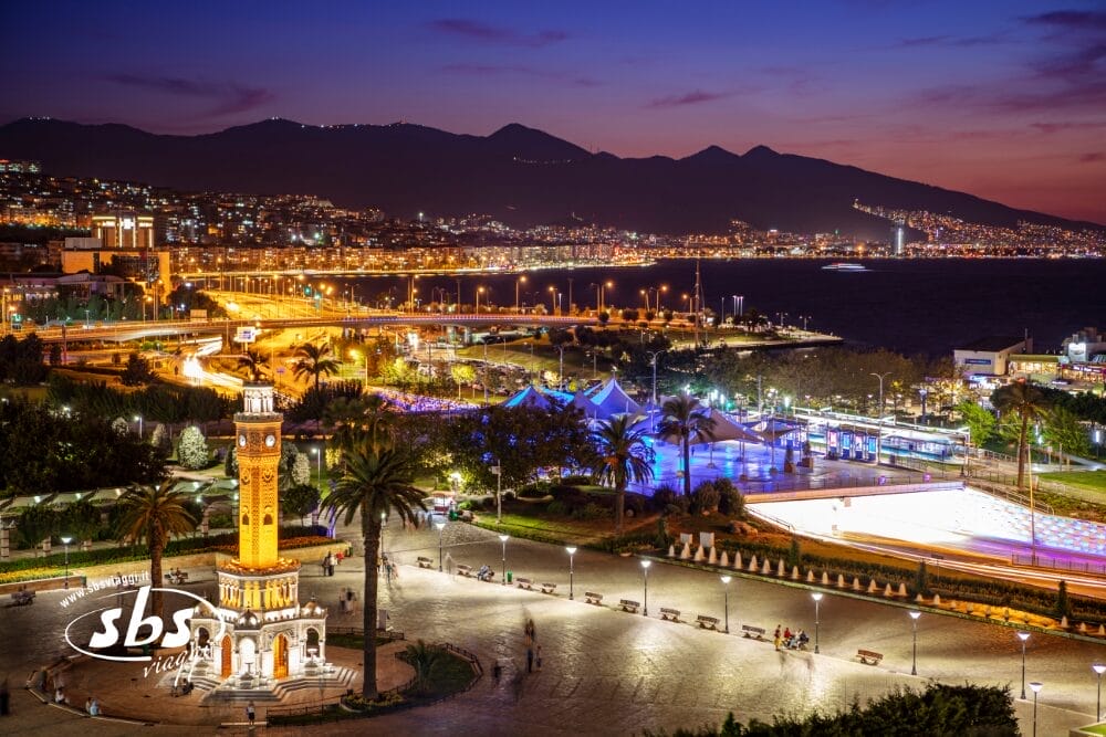 Un vibrante paesaggio urbano serale di Izmir, in Turchia, con la Torre dell'Orologio di İzmir illuminata in primo piano - una scena esclusiva di Fantasia d'Oriente con palme, luci colorate della città e il Mar Egeo contro montagne lontane.