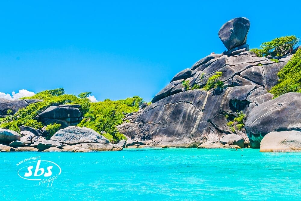 Gigantesche rocce di granito e una rigogliosa vegetazione circondano l'acqua limpida e turchese sotto un cielo blu brillante in un'isola tropicale. Un grande masso dalla forma unica si trova in cima alle rocce, mentre la scritta "Bozza automatica" e il logo bianco "sbs" sono visibili nell'acqua.
