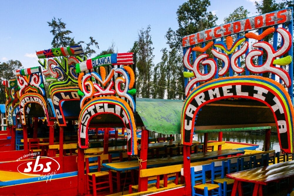 Lungo un canale, circondato da alti alberi, sono attraccate colorate trajineras messicane con insegne ornate e sedili luminosi. L'insegna della barca principale, che ricorda le scene di festa dello Yucatán o del Chiapas, recita "Carmelita" in colori vivaci.