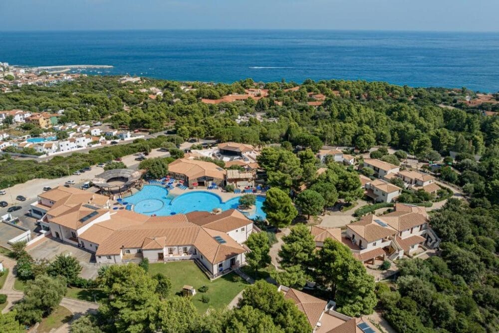 Veduta aerea di un resort costiero con edifici dai tetti abbronzati, piscine e alberi verdeggianti, rappresentato come una Bozza automatica vicino alla costa con il mare blu scintillante sullo sfondo.