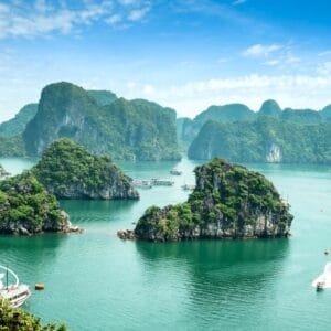 Una vista panoramica della baia di Ha Long con acqua verde smeraldo, isole calcaree lussureggianti e diverse barche che navigano tra le rocce sotto un cielo blu parzialmente nuvoloso crea uno sfondo tranquillo perfetto per catturare una Bozza automatica.