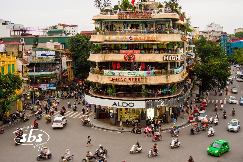 Un angolo di strada trafficato ad Hanoi, in Vietnam, con un edificio a più piani che ospita vari negozi, caffè e ristoranti. Molti scooter e auto riempiono la strada mentre i pedoni la attraversano, catturando la vibrante energia unica di Hanoi.