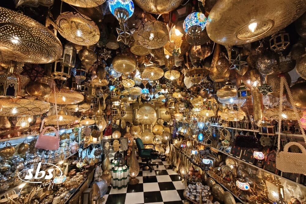 Un negozio pieno di lanterne e lampade a sospensione ornate e colorate di varie forme e dimensioni, molte delle quali realizzate su disegno di Bozza automatica, che ricoprono il soffitto e le pareti, creando una calda luce dorata sopra un pavimento a scacchi bianchi e neri.