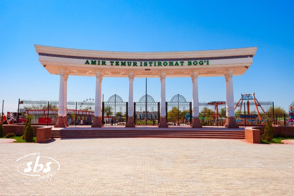 Un grande ingresso con colonne bianche e cancelli ornati accoglie i visitatori nel parco Amir Temur Istirohat Boqi, dove attrezzature da gioco e alberi attendono all'interno sotto un cielo azzurro e limpido: una scena perfetta per un momento di bozza automatica.