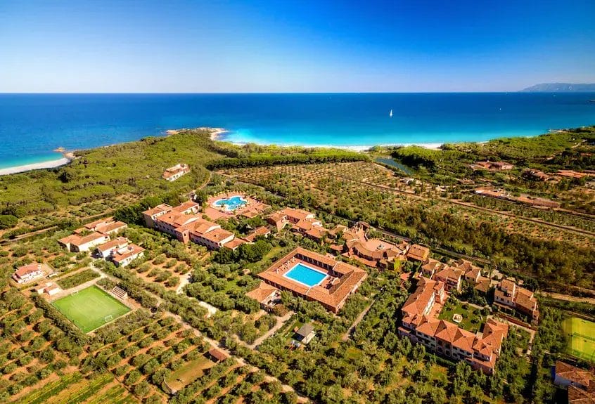 Vista aerea di un resort costiero immerso nel verde, con edifici dai tetti rossi, due piscine, campi da tennis e una spiaggia sabbiosa che incontra il mare blu sotto un cielo limpido: un rifugio idilliaco pronto per la vostra prossima Bozza automatica.