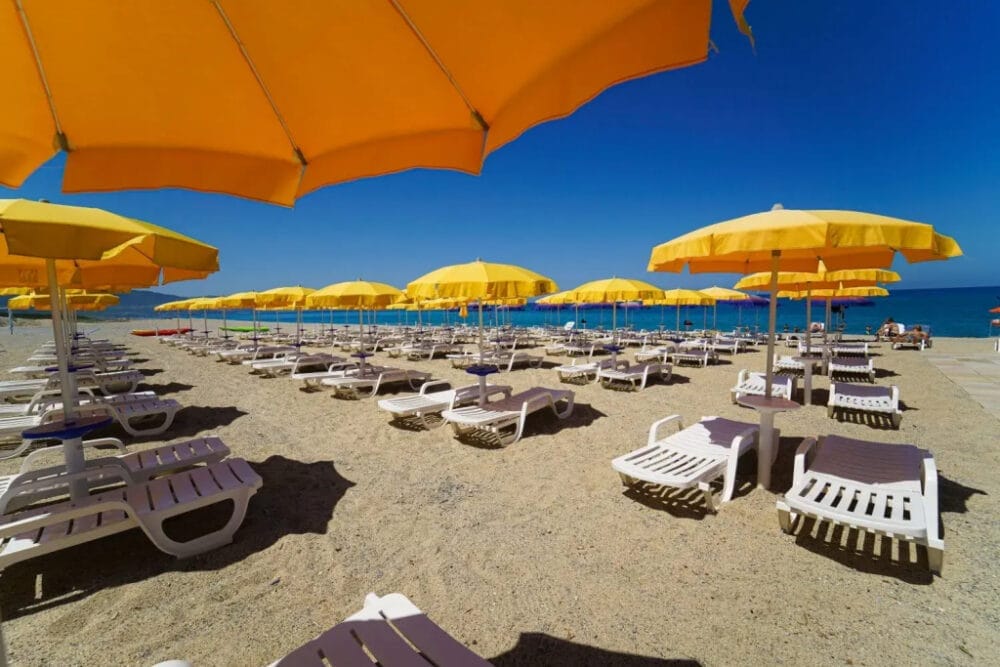 File di sedie a sdraio bianche vuote e ombrelloni gialli costeggiano la spiaggia sabbiosa sotto un cielo azzurro e limpido, con l'oceano sullo sfondo: una scena invitante per una giornata di relax, catturata prima dell'arrivo degli ospiti, quasi come una bozza automatica.