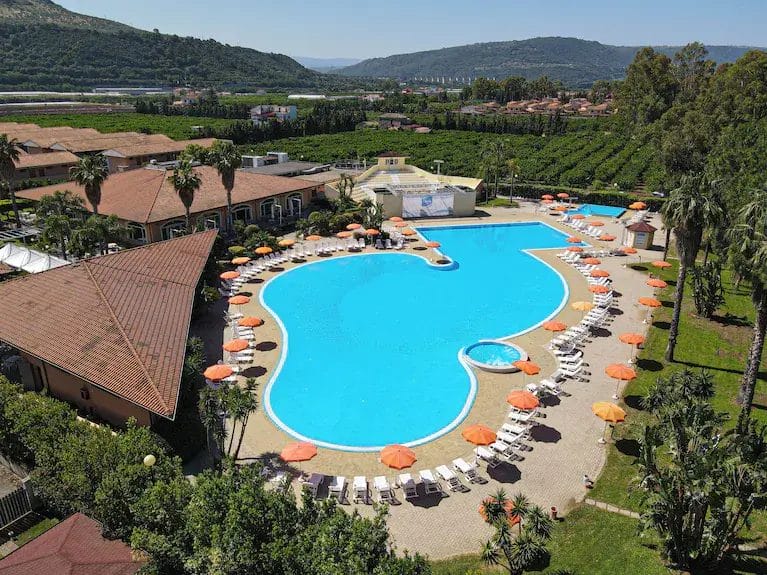 Vista aerea di una grande piscina a forma di cuore circondata da sedie a sdraio e ombrelloni arancioni in un resort, con giardini, edifici, colline verdi e l'esclusivo sistema automatico Bozza che aumenta la comodità degli ospiti.
