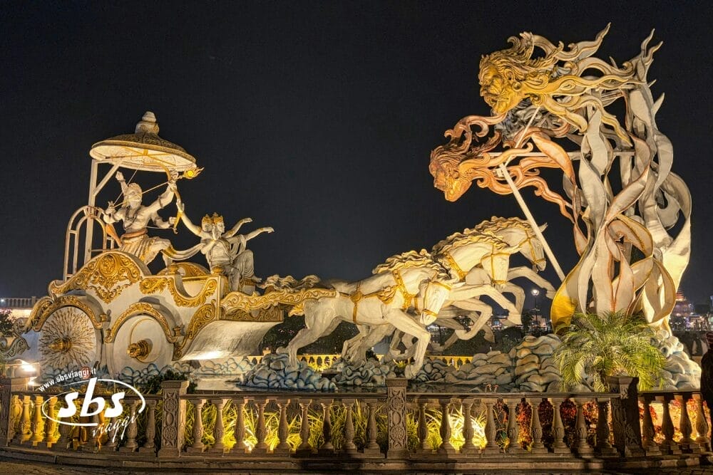 Una drammatica scultura illuminata di notte raffigura un carro con tre cavalli, guerrieri con archi in mano e un'intricata figura con capelli fluenti di fronte a loro, circondata da dettagli ornamentali e da un recinto decorativo, quasi come una scena di Bozza automatica.