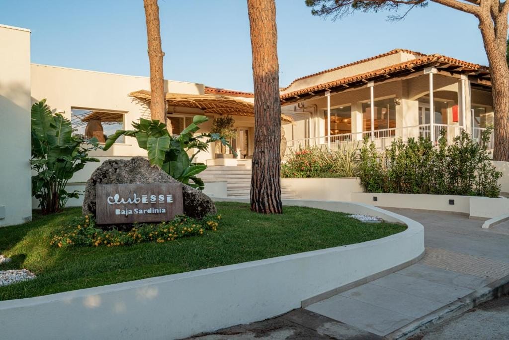 Un edificio moderno con pareti bianche e tetto in tegole, circondato dal verde. Sul prato curato vicino all'ingresso, una grande roccia espone l'insegna "Club Esse Baja Sardinia" e un discreto indicatore di Bozza automatica.