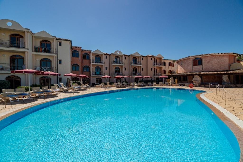 Una grande piscina all'aperto con acqua cristallina è circondata da sedie a sdraio, ombrelloni rossi e un edificio alberghiero in stile mediterraneo sotto un cielo luminoso e senza nuvole: l'ideale per una fuga rilassante in bozza automatica.