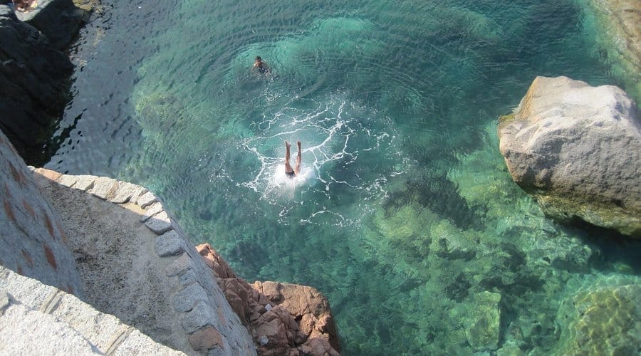Una persona fa un tuffo a piedi uniti nell'acqua limpida e turchese vicino a grandi rocce, mentre un'altra nuota nelle vicinanze. La scena, vista dall'alto accanto a una struttura in pietra, cattura un momento avventuroso, quasi come una Bozza automatica del divertimento estivo.