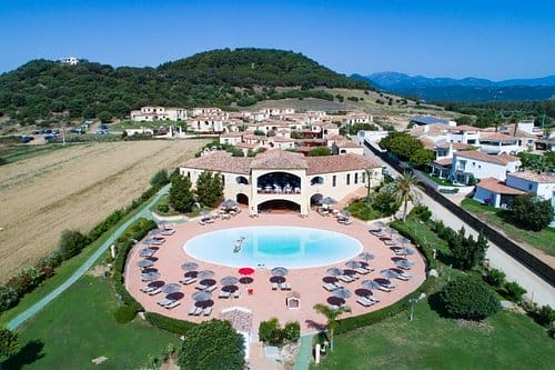 Vista aerea di una grande piscina circolare circondata da sedie a sdraio e ombrelloni, immersa in un'area paesaggistica con edifici e colline vicine sullo sfondo, sotto un cielo azzurro e limpido: un rifugio ideale per Bozza automatica.