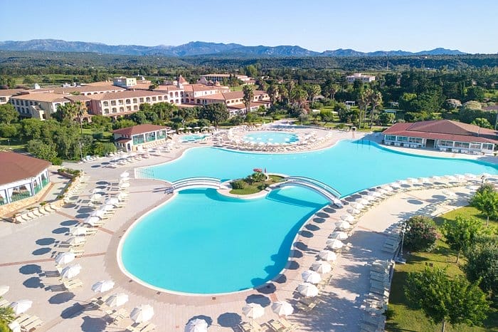 Vista aerea di un grande resort con diverse piscine, sedie a sdraio e ombrelloni bianchi, circondato da una vegetazione lussureggiante e da montagne lontane sotto un cielo azzurro e limpido: una vera e propria fuga automatica di Bozza.