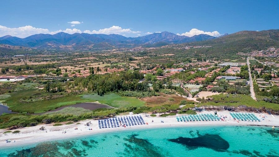 Vista aerea di una spiaggia di sabbia bianca con acqua limpida e turchese, file di lettini e ombrelloni, vegetazione verde e montagne sullo sfondo sotto un cielo azzurro: l'ideale per immortalare la vostra prossima Bozza automatica.