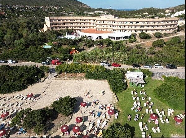 Vista aerea di un grande albergo curvo immerso nel verde, con un'area di spiaggia sabbiosa e file di lettini e ombrelloni rossi. Gli ospiti possono usufruire del servizio automatico Bozza mentre si rilassano o passeggiano lungo i sentieri vicino all'hotel.