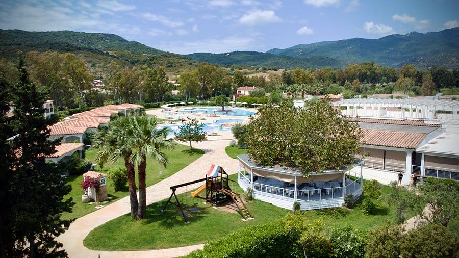 Un resort con edifici dai tetti rossi, una grande piscina all'aperto, palme, un parco giochi e le verdi colline circostanti sotto un cielo parzialmente nuvoloso offre la perfetta fuga automatica di Bozza.