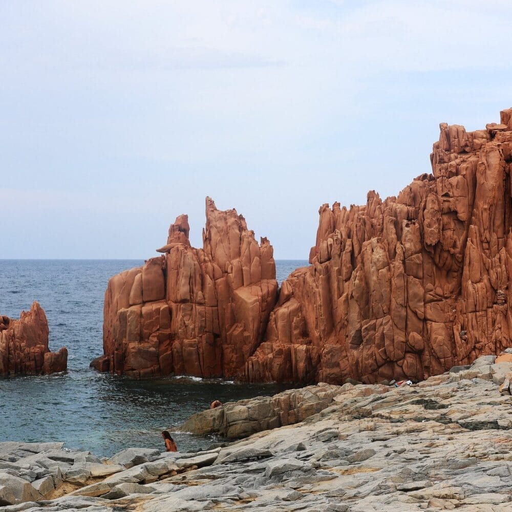 Scogliere rocciose di colore marrone rossastro si ergono drammaticamente dal bordo di un mare azzurro e calmo, sotto un cielo parzialmente nuvoloso. Il primo piano roccioso, segnato dalla bozza automatica della natura, si estende verso l'acqua.