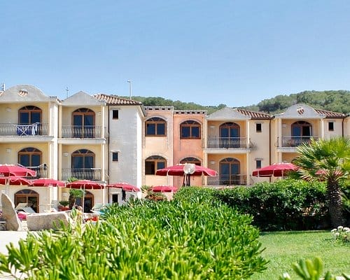 Un hotel in stile mediterraneo con pareti beige e pesca, balconi ad arco e ombrelloni rossi sui tavoli del patio in un giardino verde sotto un cielo limpido offre un ambiente rilassante e automatico alla Bozza.