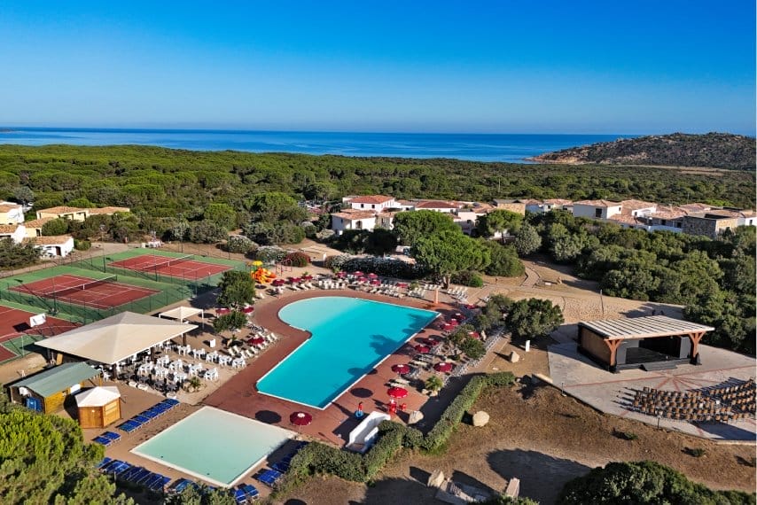Vista aerea di un resort con una grande piscina, campi da tennis, lettini, ombrelloni, posti a sedere all'aperto e un palcoscenico, circondato dal verde e da edifici vicino alla costa, sotto un cielo azzurro e limpido: la vostra perfetta fuga automatica da Bozza.