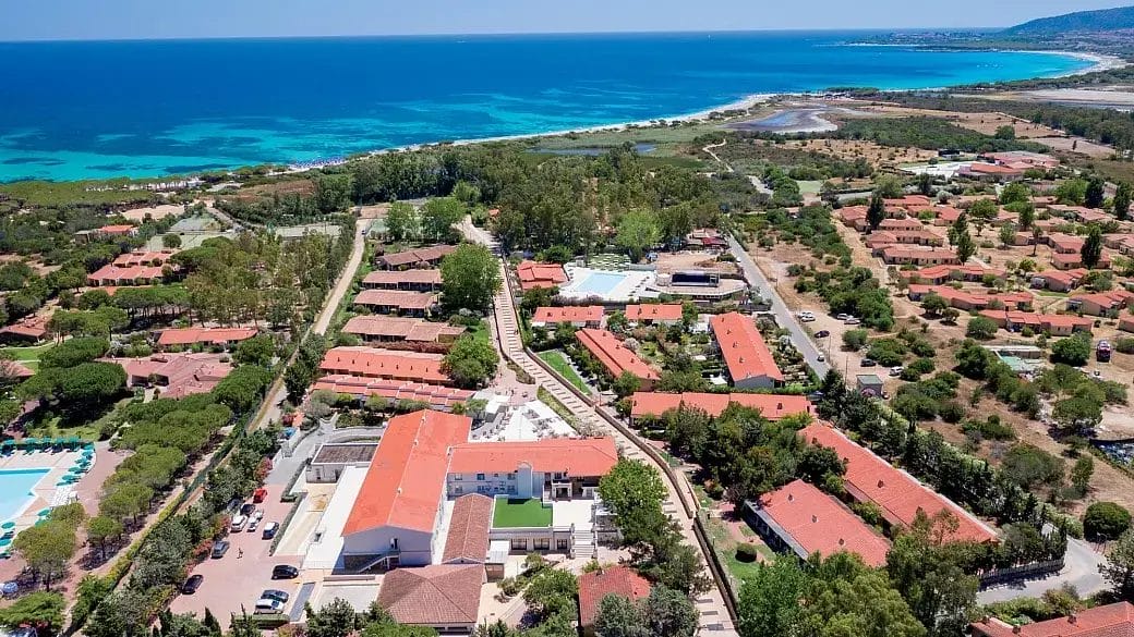 Vista aerea di una località balneare con edifici dai tetti rossi, alberi verdi e una costa turchese sotto un cielo azzurro e limpido. Questa Bozza automatica cattura l'oceano che si estende lungo spiagge sabbiose e vegetazione lussureggiante.