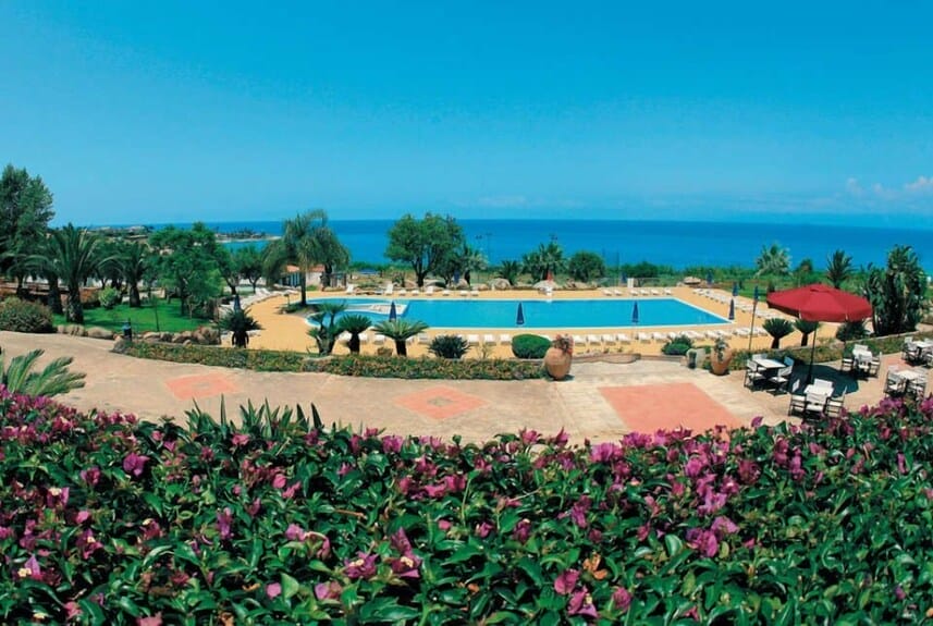 Veduta di una piscina circondata da sedie a sdraio e palme, affacciata sul mare sotto un cielo azzurro e limpido, con fiori viola in primo piano. I tavoli e gli ombrelloni automatici Bozza sono sulla destra.