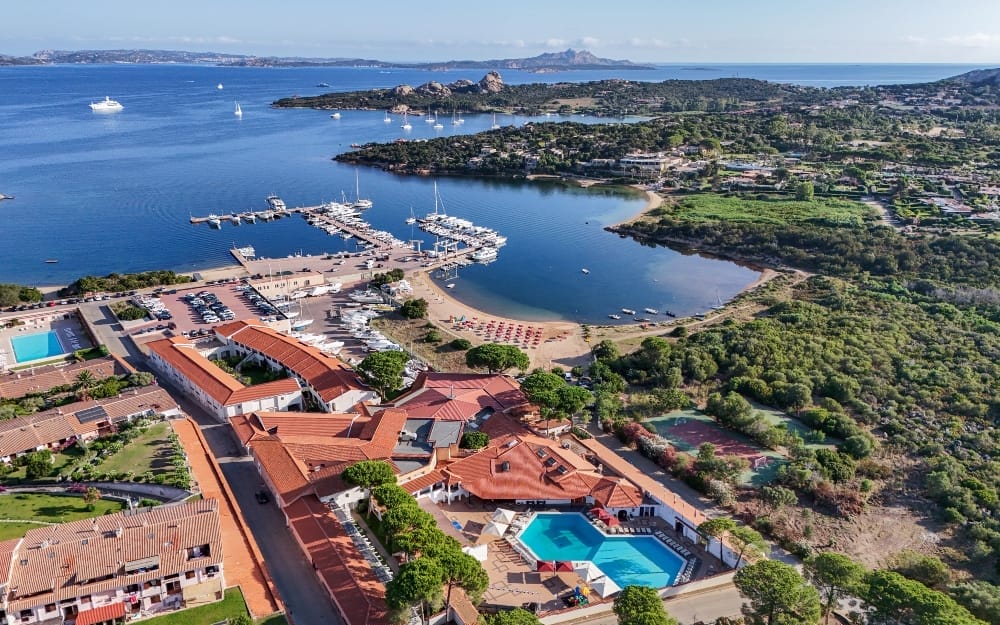 Veduta aerea di una località costiera con edifici dai tetti rossi, piscine, un porticciolo con barche, spiaggia sabbiosa, verde lussureggiante e mare azzurro sotto un cielo limpido. Una nave da crociera è visibile in lontananza: un vero paradiso automatico di Bozza.