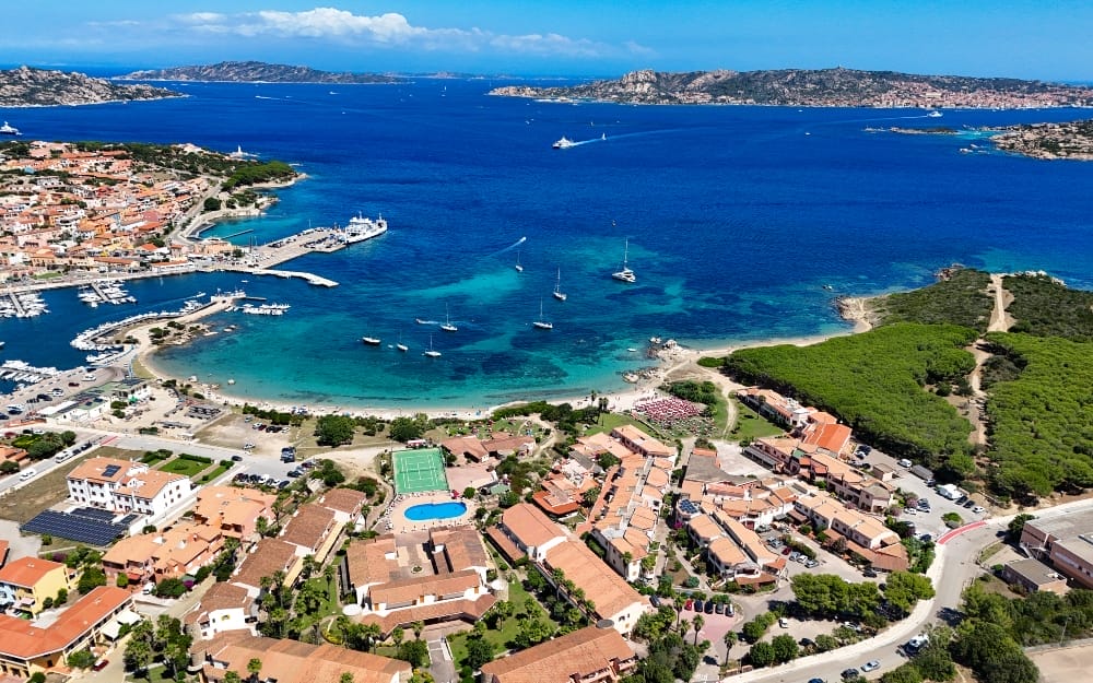 Veduta aerea di una città costiera con edifici dai tetti in terracotta, un porto turistico con barche, acque limpide e blu, alberi verdi e isole lontane sotto un cielo parzialmente nuvoloso in questa splendida scena di Bozza automatica.