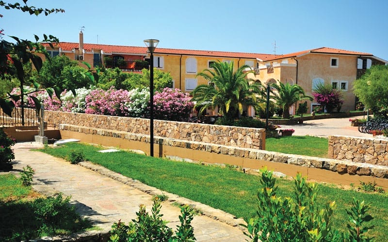 Un cortile soleggiato con erba verde, cespugli fioriti, palme e un muro di pietra sembra una bozza automatica di fascino mediterraneo, incorniciata da edifici con tetti in terracotta e pareti giallo caldo sullo sfondo.