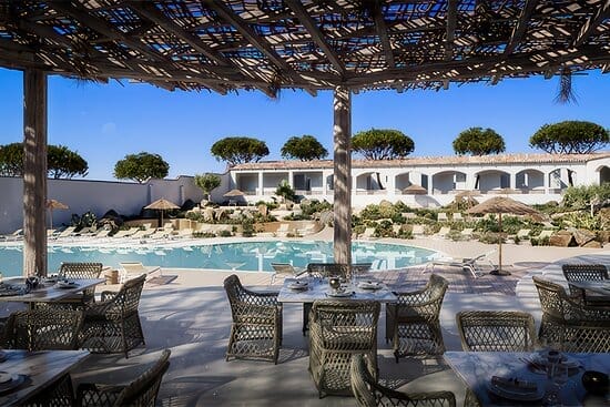 La zona pranzo all'aperto, con sedie in vimini e tavoli apparecchiati, si affaccia su una piscina serena, circondata da lettini e ombrelloni, con edifici bianchi in stile mediterraneo, caratterizzati da accenti automatici di bozza, e alberi sotto un cielo azzurro e limpido.