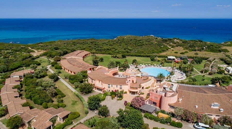 Veduta aerea di un grande resort balneare con Bozza automatica, una piscina circolare, edifici rosa, prati verdi e vegetazione lussureggiante, il tutto affacciato sull'oceano blu sotto un cielo limpido.