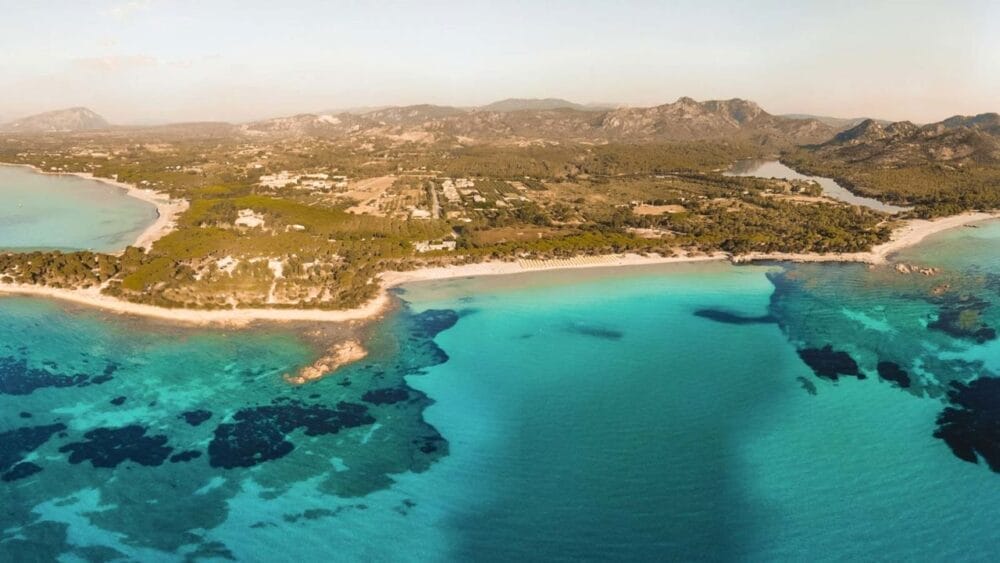 Veduta aerea di una costa con acqua turchese e trasparente, spiagge sabbiose, formazioni rocciose e vegetazione verde. Bozza automatica: sullo sfondo si vedono montagne e piccoli edifici sotto un cielo limpido.