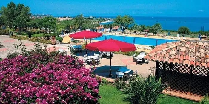 Un resort panoramico con piscina, tavoli ombreggiati da ombrelloni rossi, giardini lussureggianti con fiori viola, palme e una vista sul mare sotto un cielo azzurro e limpido: l'ideale per rilassarsi o per preparare la prossima bozza automatica.