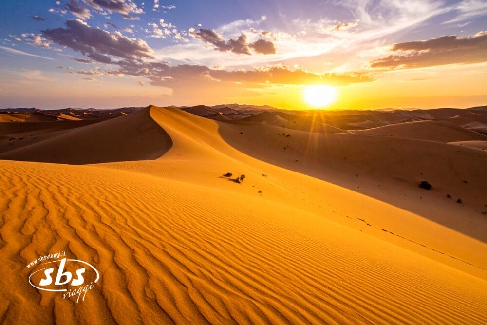 Dune di sabbia dorata si estendono in lontananza sotto un cielo drammatico al tramonto, perfetto per una Fuga nel Deserto. Il sole proietta lunghe ombre sul paesaggio, con il logo "sbs viaggi" nell'angolo in basso a sinistra.