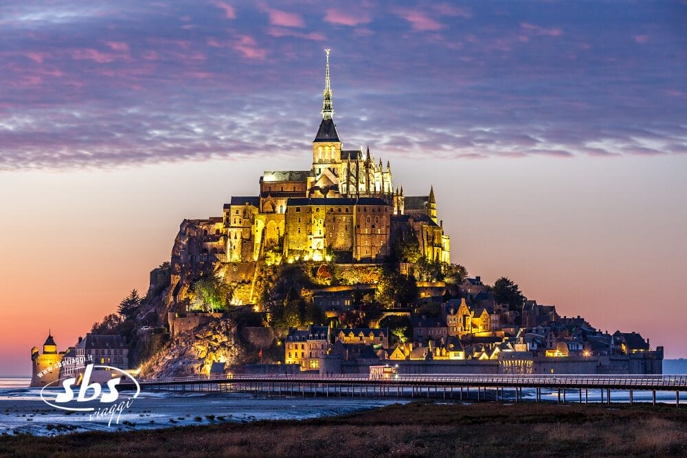 Mont Saint-Michel in Normandia illuminato al tramonto, con la sua storica abbazia e gli edifici circostanti che brillano sotto il cielo colorato della sera, e il lungo ponte che conduce all'isola.