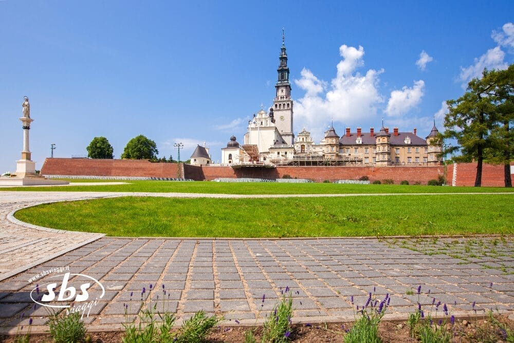 Una veduta del monastero di Jasna Góra a Częstochowa, in Polonia, con la sua alta torre e gli edifici storici si staglia come una bozza automatica, incorniciata da prati verdi, alberi e una statua in primo piano sotto un cielo azzurro.