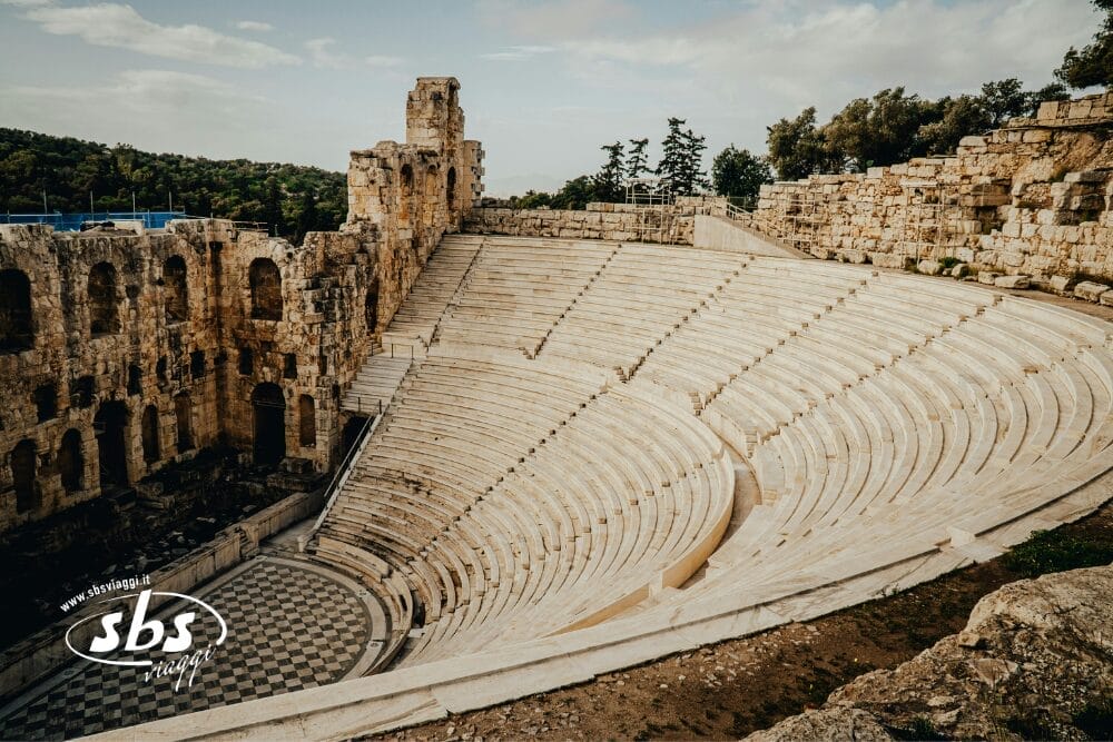 Antico anfiteatro in pietra con gradinate e un muro parzialmente rovinato, circondato da alberi e da un cielo nuvoloso. Il pavimento dell'arena presenta un disegno. Nell'angolo in basso a sinistra compaiono le scritte "Bozza automatica", "sbs" e il sito web.