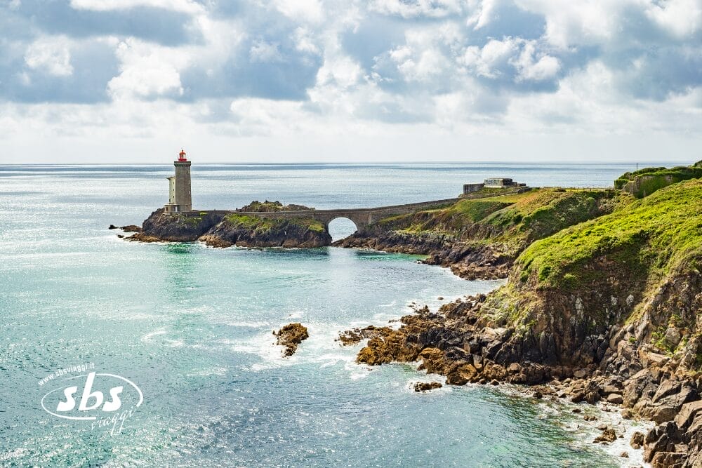 Un faro si trova alla fine di un ponte di pietra su una costa rocciosa in Normandia, circondato da un mare verde-azzurro e da scogliere verdi sotto un cielo parzialmente nuvoloso.