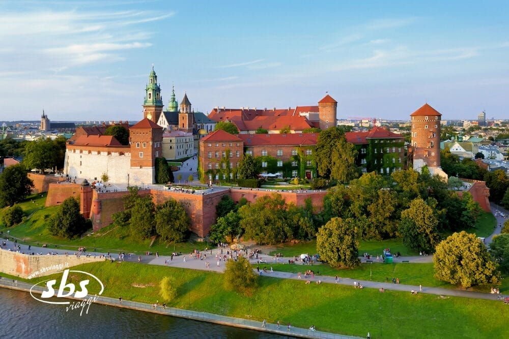 Vista aerea del Castello di Wawel a Cracovia, in Polonia, con i suoi tetti rossi, le torri e il verde circostante lungo il fiume Vistola. Le persone si godono il parco sottostante in una giornata di sole, ideale per immortalare una Bozza automatica.