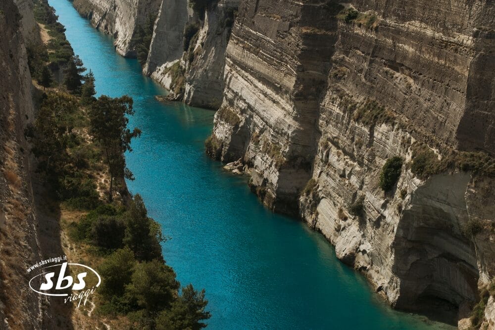 Uno stretto canale blu turchese scorre tra ripide scogliere rocciose con una rada vegetazione ai margini sotto la luce del giorno. Il logo di SBS Viaggi e la frase "Bozza automatica" appaiono nell'angolo in basso a sinistra.