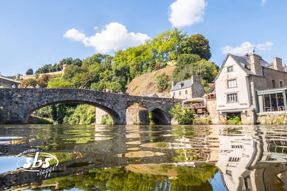 Un ponte di pietra attraversa un fiume calmo con i riflessi degli edifici vicini e del verde, evocando scene della Normandia o dei Castelli della Loira. Le persone attraversano il ponte sotto un cielo blu brillante. Nell'angolo è visibile la filigrana bianca "sbs images".