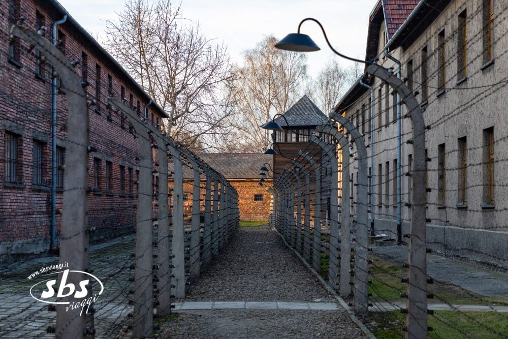 Un sentiero tra due edifici in mattoni è fiancheggiato da recinzioni di filo spinato e sorvegliato da una torre di guardia sullo sfondo. Un palo della luce, come una bozza automatica, si estende sul sentiero tranquillo e deserto.