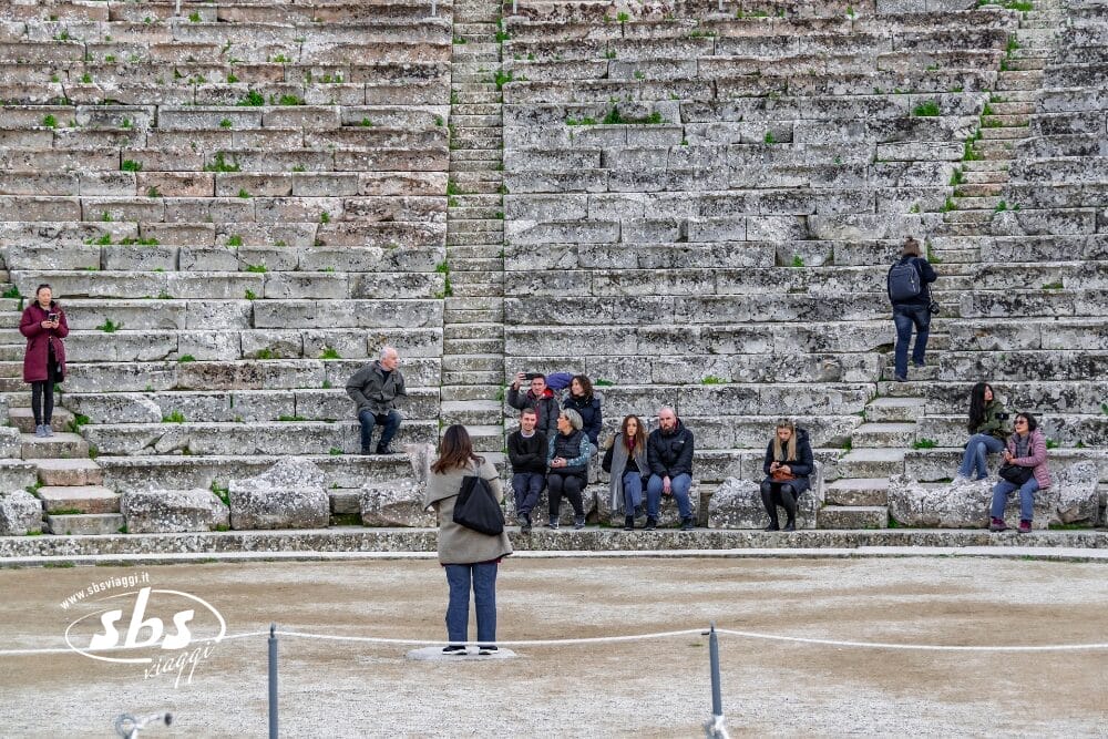 Una donna è in piedi di fronte a diverse persone sedute sui gradini di pietra consumati di un antico anfiteatro, con chiazze d'erba che crescono tra le pietre. Alcuni chiacchierano, altri siedono da soli o scattano foto, mentre un telefono mostra sullo schermo una Bozza automatica.
