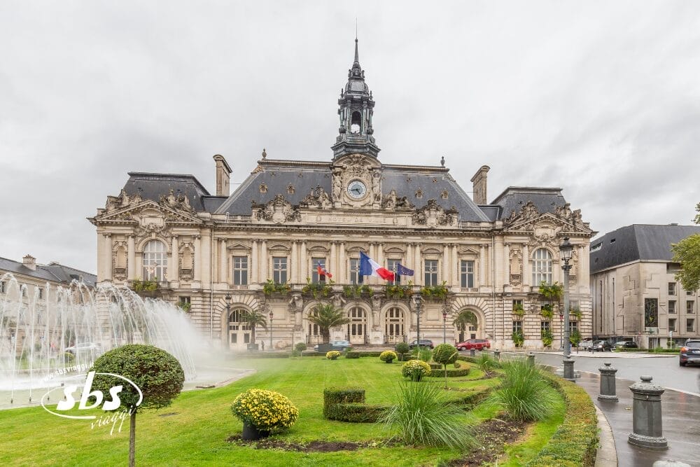 Un grande edificio storico con architettura ornata, torre dell'orologio, bandiere francesi e finestre ad arco si erge dietro un giardino curato che ricorda i Castelli della Loira, ornato da fontane e arbusti, sotto un cielo nuvoloso di Parigi.