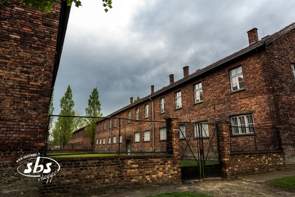 Veduta di edifici in mattoni dietro un cancello e una recinzione metallica del campo di concentramento di Auschwitz, con il cielo nuvoloso e alcuni alberi visibili sullo sfondo. L'immagine include la filigrana "Bozza automatica" e recita "sbs.