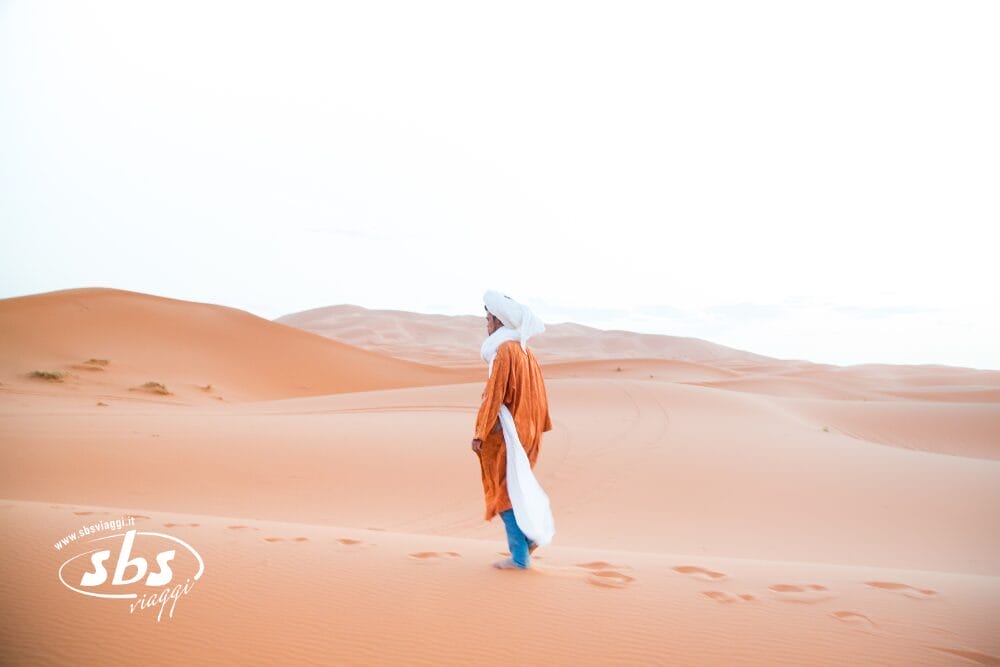 Una persona con un turbante bianco e una tunica arancione cammina da sola attraverso morbide dune di sabbia sotto un cielo limpido e pallido, lasciando dietro di sé delle impronte: un vero e proprio viaggio deserto attraverso un paesaggio vasto e aperto.