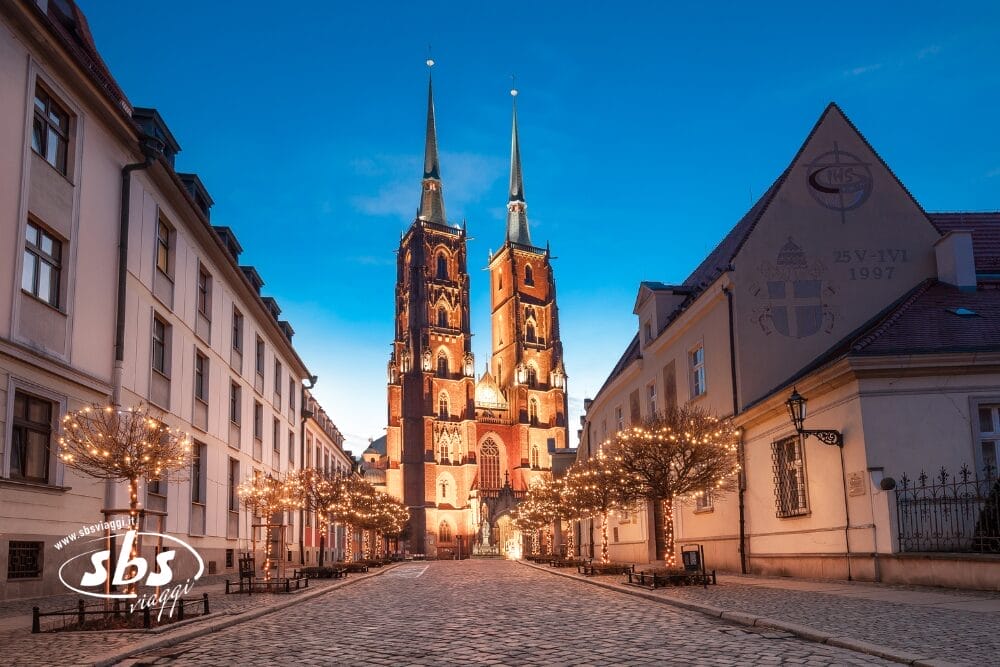 Una strada di ciottoli fiancheggiata da alberi illuminati conduce a una grande cattedrale gotica illuminata con due alte guglie, circondata da edifici storici all'imbrunire: una bozza automatica di fascino antico e serenità serale.