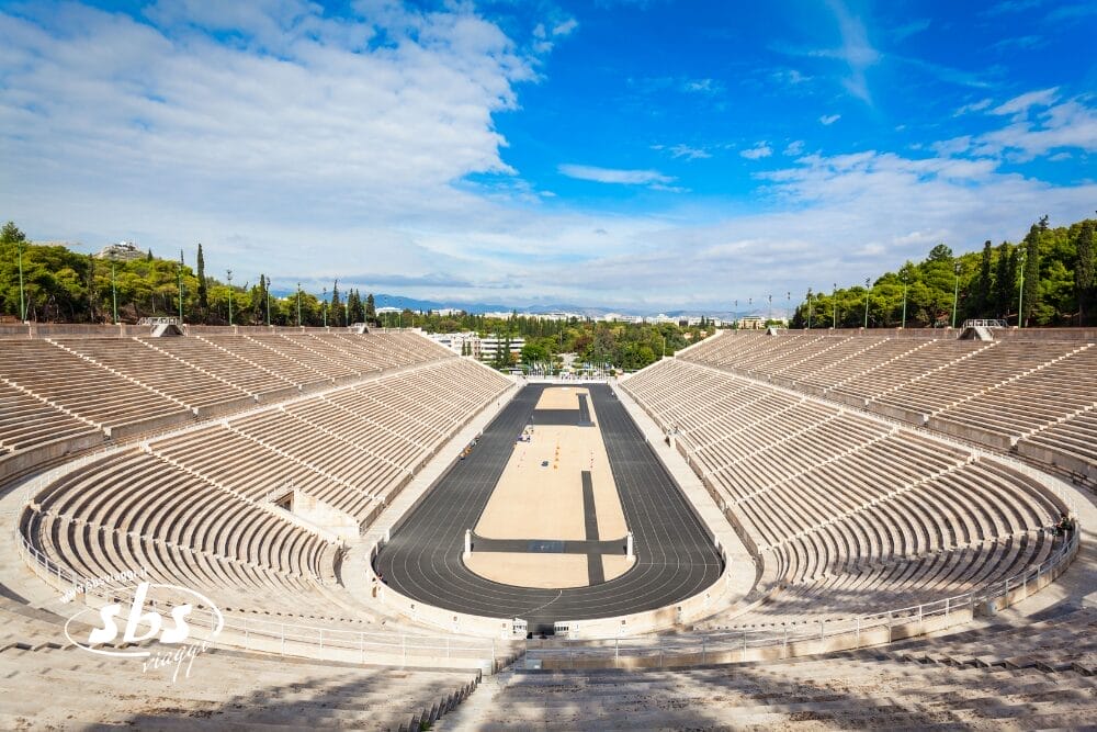 Vista panoramica dello Stadio Panathenaic di Atene, in Grecia, che mostra i posti a sedere in pietra vuoti e la pista di corsa centrale - un'impressionante Bozza automatica contro gli alberi e un cielo parzialmente nuvoloso, con alcuni visitatori che camminano.