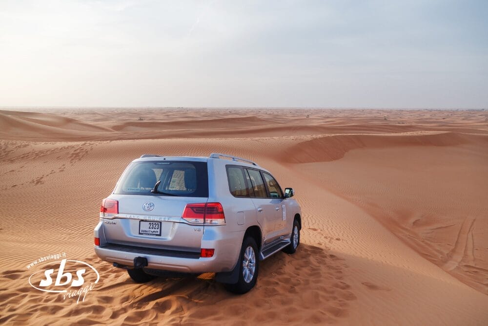 Un SUV argentato è parcheggiato su una duna sabbiosa del deserto sotto un cielo limpido, che mostra le tracce di pneumatici nella sabbia e le vaste dune al di là - una scena perfetta per una Fuga nel Deserto o un emozionante tour di 5 giorni nel deserto.