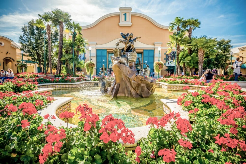 Un giardino vivace con fiori rossi circonda una fontana decorativa con una statua di bronzo, situata vicino all'ingresso di Disneyland Paris, incorniciata da palme e da un edificio giallo sotto il cielo blu.